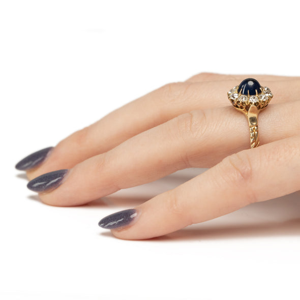 Victorian 14K Yellow Gold Deep Blue Cambodian Cabochon Sapphire and Old European Cut Diamond Halo Cocktail Ring on the hand from the side showcasing the sugar loaf cab sapphire 